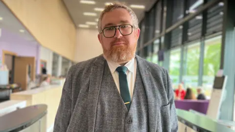 BBC/Aisha Iqbal Edwards standing indoors and positioned centrally in the foreground. He is wearing rectangular glasses with dark frames and has short hair along with a full beard.
The clothing consists of a grey, check-patterned suit jacket layered over a matching waistcoat and a white collared shirt. A dark green tie with a tie clip is neatly placed at the centre of the outfit.