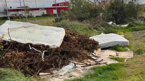 Mike Sib Multiple mattresses are piled on to sticks, plastic bags and wet cardboard in a field. A KFC building can be seen in the distance.