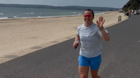 Tegan Pryce is waving to the camera as she runs along a road which runs parallel to an empty beach on a sunny day. She is wearing pale blue running shorts, a white T-shirt and a pair of sunglasses.
