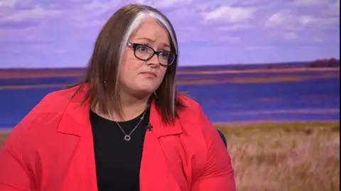 A woman with brown shoulder length hair, and two white stripes down the front of her hair. She wears glasses and a black jumper, with a red shite over it. She is in a BBC studio and looks off camera towards the presenter