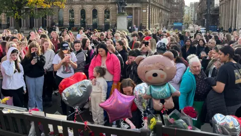 Linda Sinclair / BBC Une foule se rassemble, certaines personnes pleurent, devant un mémorial avec des ballons sur une place de Glasgow