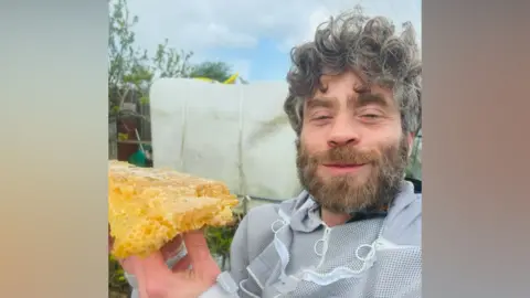 Nathan Wylam A man with curly greying brown hair and a beard smiles as he holds honeycomb up. He is wearing a beekeeping suit without the hood.
