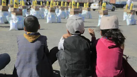 Three children sitting, with their backs to the camera, facing bags of aid