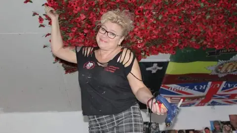 Julie Cooper Julie Cooper placing red poppies on the ceiling. She's got blonde curly hair and is holding a glue gun.