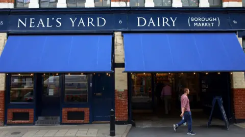 Getty Images «Neil's Yard Dairy» написано белым и синим цветом на внешней стороне магазина на рынке Боро, с пустыми полками в переднем окне и мужчиной в красной рубашке и синих джинсах, идущим справа от магазина.