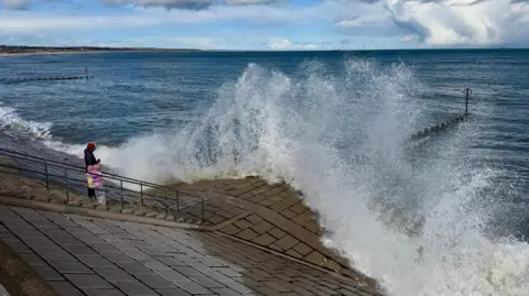 BBC Weather Watchers/clairewn A big wave hits brickwork at the shore as an adult and child watch on, wrapped up in layered warm clothing