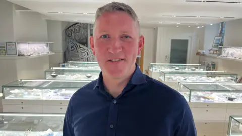 Martin is a grey-haired man in a dark blue open-necked shirt. He is standing in the middle of a jewellery gallery with glass cases behind him.