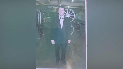 Michael Denner An old photograph of Michael in black tie standing smiling to camera in a windowless room with a large two-reel projector behind him 