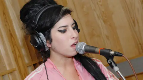 Amy Winehouse, with black hair and black eyeliner, wears a pink Adidas tracksuit and yellow vest - she is singing into a microphone, in a wooded studio 