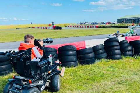 Claire Smith Jake Smith, a young man in a wheelchair, photographed from the side, he is holding a camera and taking motorsport photos at a race circuit.