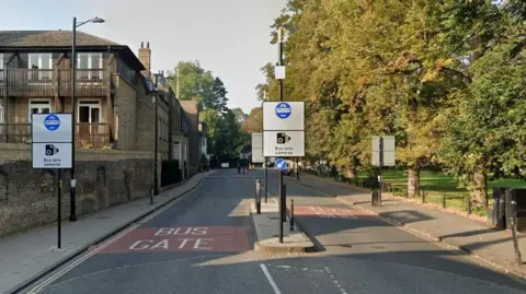 Google Photo of a bus door on Silver street in Cambridge. There are signs showing which vehicles can go on the road. There is a park on the right of the road and buildings on the left.