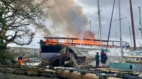 The boat is burning and there are many flames and much smoke. Three people are watching the spectacle. It's a cloudy day and there is a tree near the boat and other boats moored next to it.