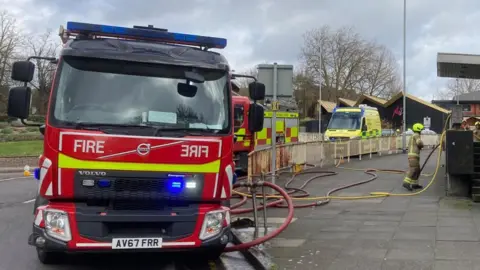 Andrew Woodger/BBC Two fire engines can be seen parked on a road with an ambulance behind. A fire officer with protective gear is on the pavement and fire hoses are too.  