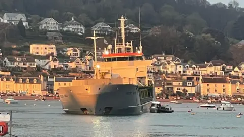 A large silver cargo ship which run aground in Teignmouth harbour during the early hours. The photo was taken during the sunrise. Several other boats are docked in the water. A pilot vessel is next to the cargo ship.