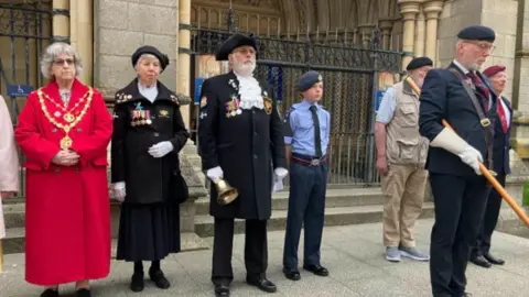 Lionel Knight Truro town crier seen wearing a black military-style uniform with a large bell in his hand. He is stood on the steps of Truro Cathedral with other people wearing uniform.
