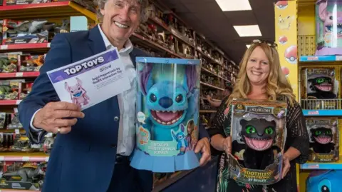 Wow! Stuff A man and a woman holding toys and smiling. Toys are on shelves in the background.
