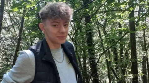 A young man with short curly brown hair is smiling while standing next to trees. He is wearing a silver chain necklace, a grey sweatshirt and a dark blue gilet.