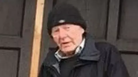 An image of Alan Lloyd. It is a blurred image, possibly from a distance and zoomed in. He is a white man, elderly, wearing a black beanie hat, black rain coat, and black jumper with a white or light-coloured shirt collar showing. He has a neutral expression.