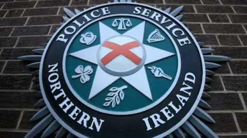 A close up of the Police Service of Northern Ireland crest against a brown brick wall