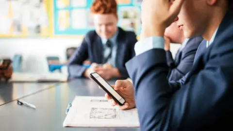 Students in school looking at their phones. We can see two students from the side in a navy school uniform and one student further away facing the camera who is blurred
