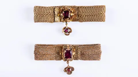 Stephen Garnett Photography Two intricately woven human‑hair bracelets with ornate gold clasps and deep red gemstones displayed side by side.