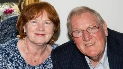 Moultrie family Pamela Moultrie has short, ginger hair and is wearing a blue and white patterned top, silver necklace and dangly earrings. Her husband Jim has short, grey hair and is wearing glasses, a black striped suit jacket and a pale blue striped shirt. They are both smiling and there is a mirror behind them.