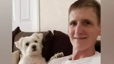 Family A man with light brown hair and wearing a white T-shirt sits on a brown sofa with a white terrier dog sitting next to him.