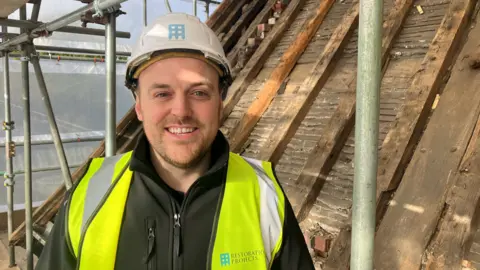 Luke Barlow on the roof of Bramall Hall in Stockport. The 36-year-old is leading the restoration project. He wears a yellow hi-vis over a dark green zipped fleece jacket.