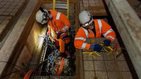 National Highways Two men wearing hard hats and orange high visibility jackets working below ground level to install cables.