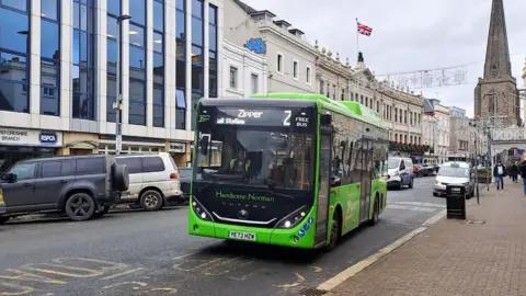 A green single decker bus parked at a bus stop in Hereford. The road has parked cars to one side with shops and office buildings to the left. In the background is a church and people walking down the nearby pavement.