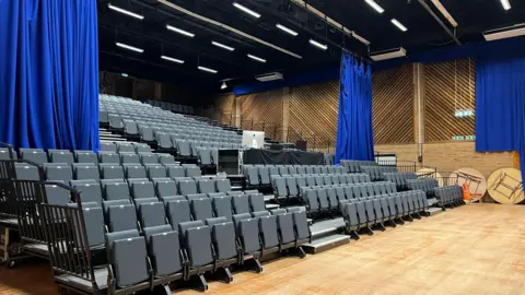 Harriet Heywood/BBC A theatre with a large wooden floor and lots of stacked tiered seating. The chairs fold down and are blue. Around the room long blue curtains are also hanging down from beams on the roof. 