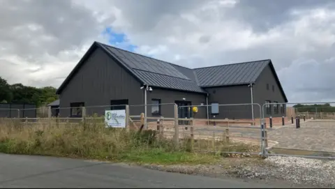 LDRS A new community centre. It is a modern building with a pitched roof. It is behind fencing. There is a paved area outside the building.