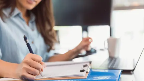Getty Images A woman sitting at a desk, writing on a clipboard with a pen. A blue folder is underneath the clipboard and a laptop and white coffee mug are visible in the background on the desk. She is wearing a light blue button-up shirt.