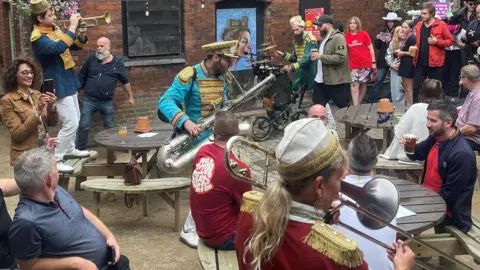 A band performing to pub goers in the Tamworth Tap Stop pub garden. The band wear blue, dark blue or burgundy jackets with gold trim on their shoulders and play brass instruments. One plays while standing on a wooden bench. The band is watched by pub goers, some who are taking footage on their phones. 
