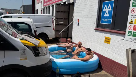 CPM BENFLEET/BBC WEATHER WATCHERS Three topless men are in an inflatable blue paddling pool, positioned outside a bricked industrial building, which is painted white and red. There are various cars and vans parked.