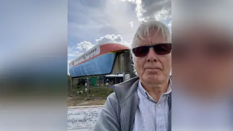 Brian Pearce Brian Pearce in a selfie photograph, wearing a fine striped shirt and grey fleece, with dark glasses. In the background is one of the original hover train models - a long carriage balancing on a concrete monorail