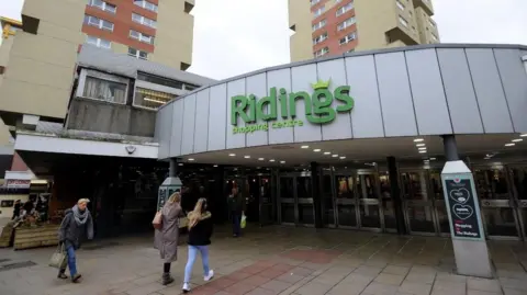 Local Democracy Reporting Service The entrance to the Ridings shopping centre. Two women walk towards a wall of glass doors set beneath a silver, grey roof. Green writing above the door says Ridings Shopping Centre. Above the shopping centre two tower blocks can be seen in the background.