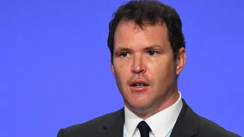 Lee Waters is standing on a stage delivering a speech. He has brown hair, is cleanly shaven. He's wearing a dark coloured suit and tie and a white shirt.