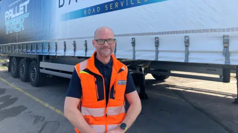Vikki Irwin/BBC Tremayne Johnson is wearing a high visibility jacket which is orange and has a Bartrums badge on the left. He is wearing a blue polo shirt underneath and is smiling at the camera. He is standing by an HGV trailer. 