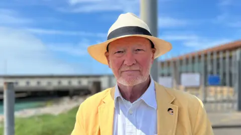 Andrew Le Quesne is standing next to Elizabeth ferry terminal, which is out of focus behind him. He is wearing a straw hat and yellow blazer and is looking into the camera.