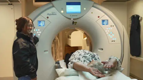 A woman lying down on a bed entering a scanning machine. A woman stands next to the machine smiling