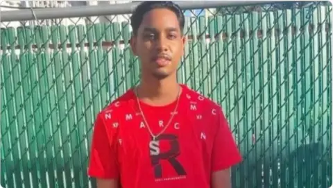 Family Shawn Seesahai wearing a red t-shirt and neck chain standing in front of green fence