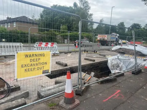A sign saying "warning deep excavation keep out" next to a fenced off area with a hole in the ground and lorries running down the A701
