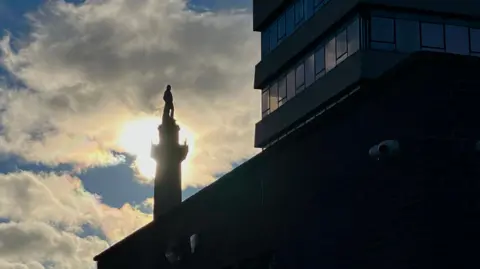 The sun breaking through clouds, forming a silhouette of a statue of a man atop a tall column, with an angular office block on the right, with sun reflecting off its banded windows