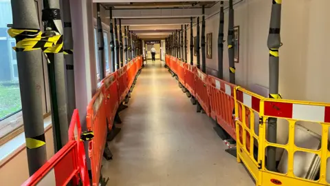 A hospital corridor is seen with yellow and orange barriers on either side, running the length of the corridor. Black and yellow tape is wrapped around pillars.