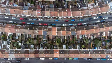 An aerial view shot of houses in Bristol. The roofs are different shades of brown and grey and cars and roads can be seen.