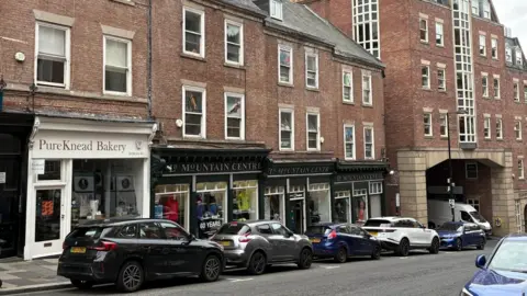 The LD Mountain Centre in Dean Street, Newcastle. The building has a shop occupying the ground floor with two floors above. Cars are parked in a row outside and Pure Knead Bakery is next door on the left.