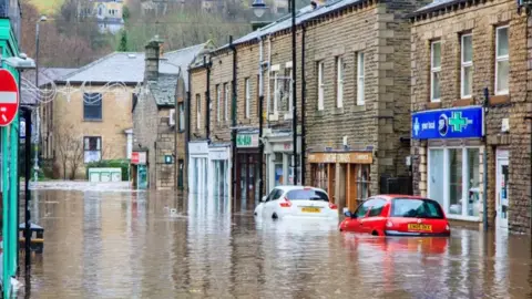 Alamy A street in Holmfirth in flood water with two partially submerged cars parked outside a pharmacy