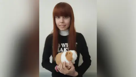 Kettering General Hospital Kirstie Pope with long red hair wearing a black top with white lettering and holding a brown and white guinea pig toy and smiling at the camera