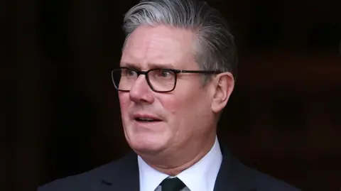 Sir Keir Starmer wearing dark rimmed glasses and looking off to his right hand side, with a dark background. He is wearing a white shirt, a dark suit and dark tie.